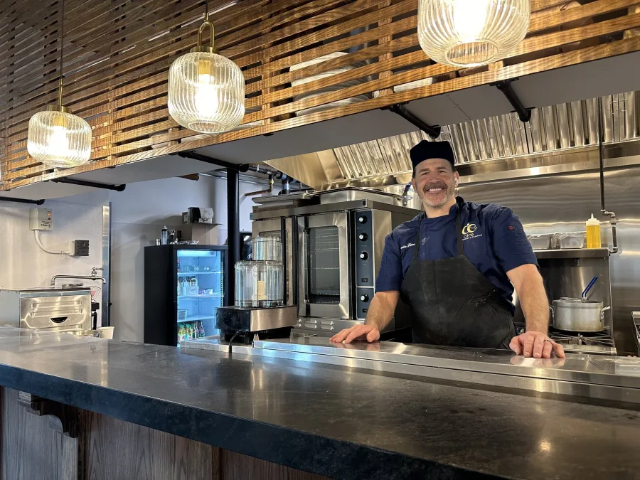 Chef smiling behind the open kitchen counter at Aire, with brass pendant lights overhead